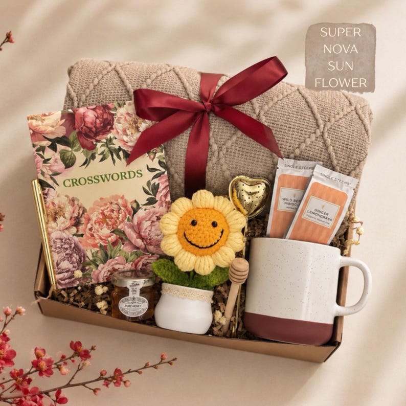 K&ouml;nnte beinhalten: Eine Geschenkbox mit einer beigen Strickdecke, die mit einem weinroten Band gebunden ist, einem floralen Kreuzwortr&auml;tselbuch, einem goldenen Stift, einem kleinen Honigglas, einer geh&auml;kelten Sonnenblume, einem herzf&ouml;rmigen Tee-Ei, zwei Teepackungen und einer Tasse.