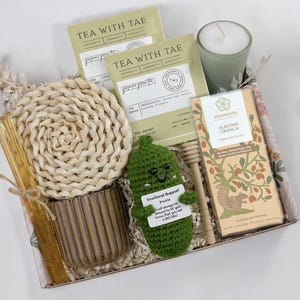 Op de afbeelding: Een cadeaudoos gevuld met verschillende items, waaronder theezakjes, een kaars, chocolade, een gehaakte augurk en een houten honingroerder. De doos is bekleed met een stof met een bloemenpatroon. De augurk heeft een bordje met de tekst "Emotional Support Pickle. I will always be there for you, no matter how bad the day is. Big Dill."