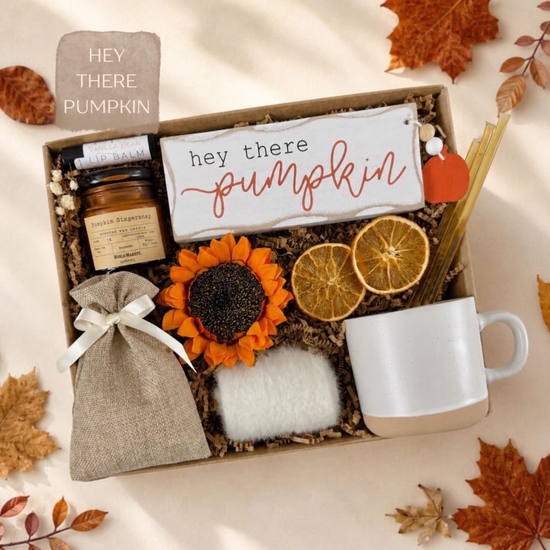 May include: A gift box containing a white sign with the text "hey there pumpkin", a small amber glass candle jar, a burlap drawstring pouch, a faux sunflower, two dried orange slices, a white ceramic mug, and a vanilla bean lip balm.