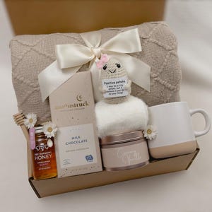 Op de afbeelding: Een cadeaupakket gevuld met zelfzorgartikelen, waaronder een pluche deken, een mok, een pot honing, een reep melkchocolade, een klein pluche aardappel speelgoed en een pot cr&egrave;me. De mand is gemaakt van karton en heeft een bruine afwerking.