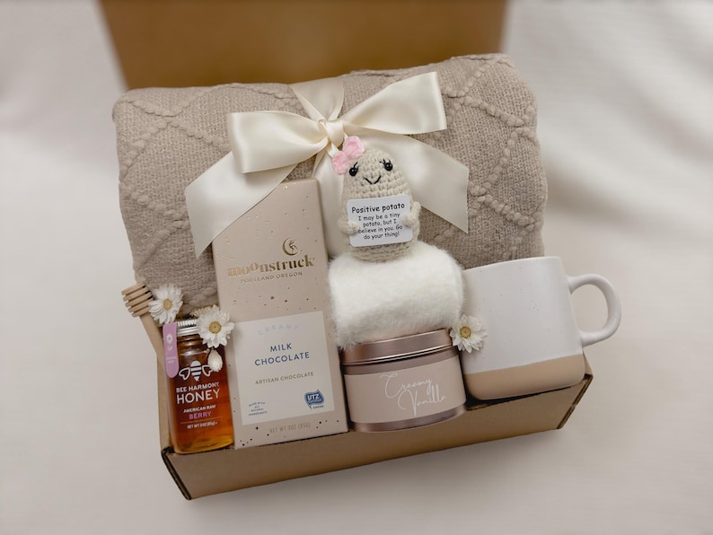 Op de afbeelding: Een cadeaupakket gevuld met zelfzorgartikelen, waaronder een pluche deken, een mok, een pot honing, een reep melkchocolade, een klein pluche aardappel speelgoed en een pot cr&egrave;me. De mand is gemaakt van karton en heeft een bruine afwerking.