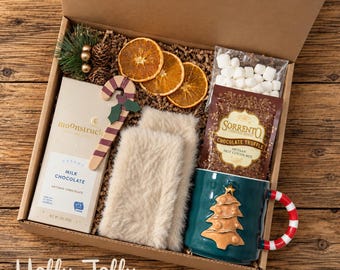 May include: A gift box containing a Moonstruck milk chocolate bar, a pair of beige faux fur mittens, a dark green mug with a gold Christmas tree design and a red and white striped handle, dried orange slices, a bag of marshmallows, and cocoa mix.