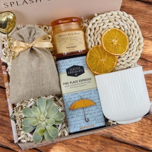 K&ouml;nnte beinhalten: Eine Geschenkbox gef&uuml;llt mit verschiedenen Artikeln, darunter eine wei&szlig;e Tasse, ein Schokoladenriegel mit dem Text "Pike Place Espresso", eine kleine Sukkulente im Topf, ein Jutesack, eine Kerze und getrocknete Orangenscheiben. Die Box ist mit braunem Papier ausgekleidet und hat ein wei&szlig;es Band.