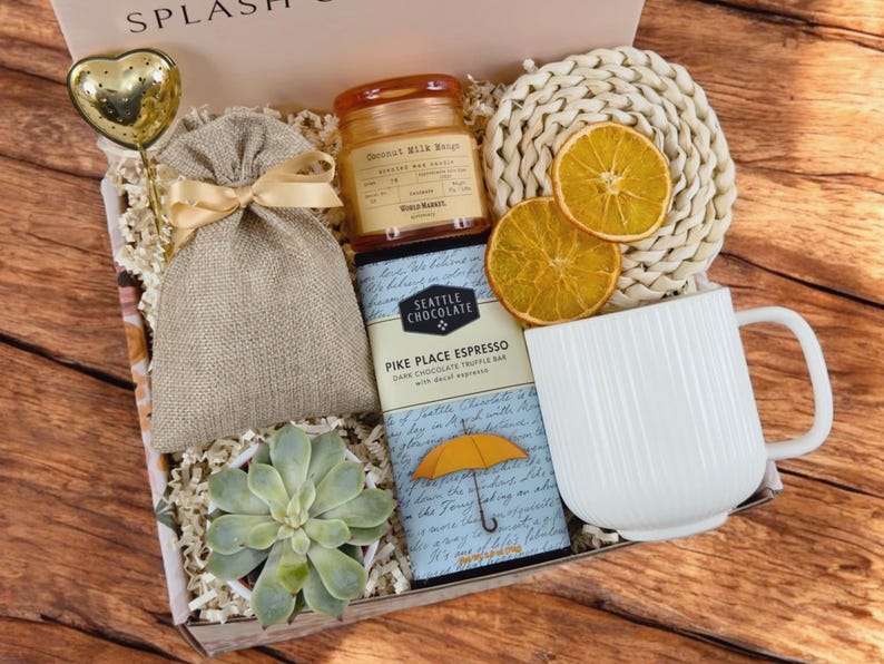 K&ouml;nnte beinhalten: Eine Geschenkbox gef&uuml;llt mit verschiedenen Artikeln, darunter eine wei&szlig;e Tasse, ein Schokoladenriegel mit dem Text "Pike Place Espresso", eine kleine Sukkulente im Topf, ein Jutesack, eine Kerze und getrocknete Orangenscheiben. Die Box ist mit braunem Papier ausgekleidet und hat ein wei&szlig;es Band.