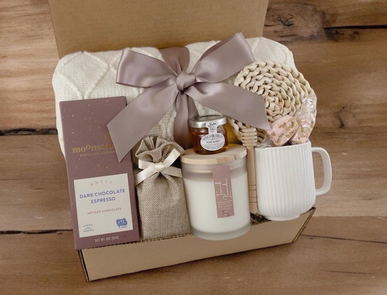 May include: A gift basket filled with self-care items, including a white knitted throw blanket, a bar of dark chocolate espresso, a candle, a jar of honey, a wooden honey dipper, a white mug, and a small burlap bag.