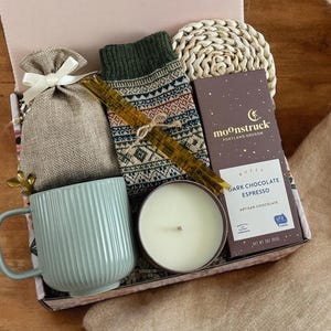 K&ouml;nnte beinhalten: Eine Geschenkbox gef&uuml;llt mit gem&uuml;tlichen Artikeln, darunter eine Tasse, eine Kerze, ein kleiner Beutel, ein gestrickter Schal und eine Tafel dunkle Schokolade mit Espresso. Die Box ist mit einem braunen und wei&szlig;en gemusterten Papier verziert und die Schokoladentafel ist mit "moonstruck, portland oregon, dark chocolate espresso, artisan chocolate" beschriftet.
