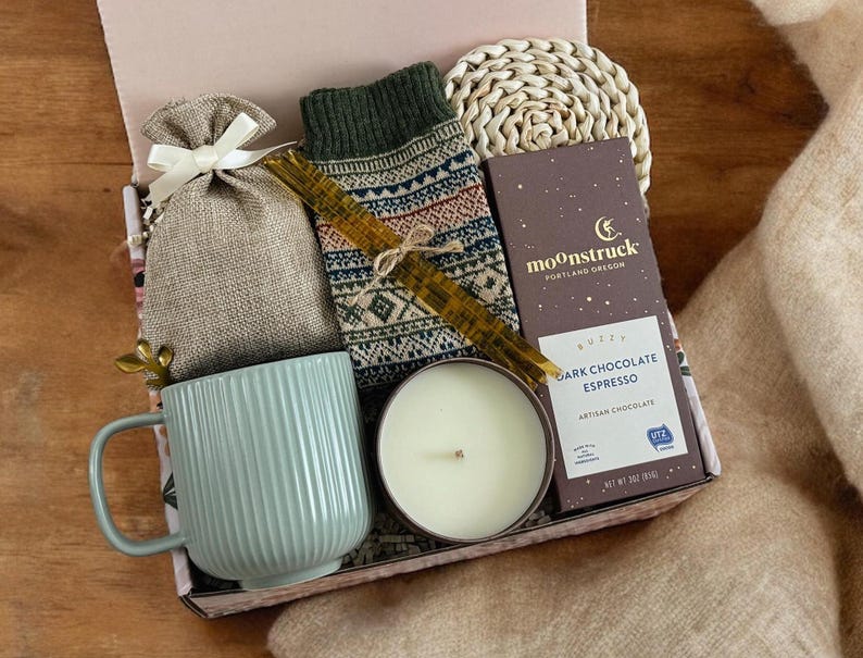 K&ouml;nnte beinhalten: Eine Geschenkbox gef&uuml;llt mit gem&uuml;tlichen Artikeln, darunter eine Tasse, eine Kerze, ein kleiner Beutel, ein gestrickter Schal und eine Tafel dunkle Schokolade mit Espresso. Die Box ist mit einem braunen und wei&szlig;en gemusterten Papier verziert und die Schokoladentafel ist mit "moonstruck, portland oregon, dark chocolate espresso, artisan chocolate" beschriftet.