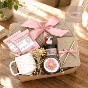 May include: A gift box labeled Blushing Daisy containing a beige knit blanket with a pink ribbon, two Tea With Tae packets, a honey jar, a black raspberry vanilla body butter, a white ribbed mug, a honey dipper, dried flowers, a candle, and a notebook with a gold pen.