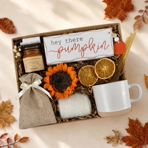 May include: A gift box filled with a white sign reading "hey there pumpkin," a small amber candle, a vanilla bean lip balm, a burlap pouch with a white ribbon, a faux sunflower, two dried orange slices, a white fuzzy item, and a white mug.
