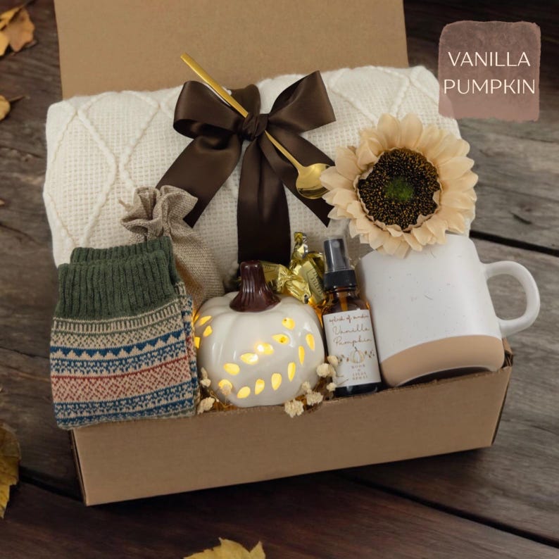 May include: A gift box containing a white cable-knit blanket tied with a brown ribbon, a sunflower, a white ceramic mug, a small white pumpkin-shaped light, a glass spray bottle labeled Vanilla Pumpkin, and patterned green knit socks.