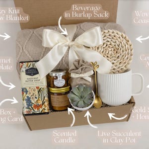 May include: A gift box filled with cozy items, including a knit throw blanket, a ceramic mug, a woven coaster, a gourmet chocolate bar, a scented candle, a live succulent in a clay pot, a beverage in a burlap sack, and a gold spoon and honey jar.