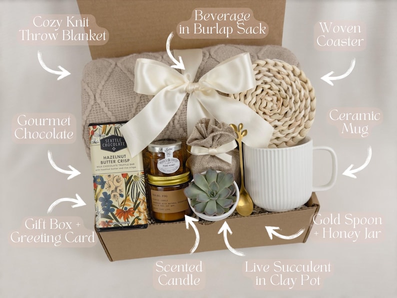 May include: A gift box filled with cozy items, including a knit throw blanket, a ceramic mug, a woven coaster, a gourmet chocolate bar, a scented candle, a live succulent in a clay pot, a beverage in a burlap sack, and a gold spoon and honey jar.
