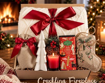 May include: A gift box containing a white cable-knit blanket tied with a red ribbon, a small burlap pouch with a red bow, a white ceramic tree, a red candle, a Seattle Chocolate Hot Cannoli bar, and a burlap stocking with embroidered greenery.