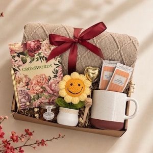 K&ouml;nnte beinhalten: Eine Geschenkbox mit einer beigen Strickdecke mit bordeauxrotem Band, einem floralen Kreuzwortr&auml;tselbuch, einem goldenen Stift, einem kleinen Honigglas, einer geh&auml;kelten Sonnenblume im wei&szlig;en Topf, einem herzf&ouml;rmigen Tee-Ei und einer Keramiktasse.