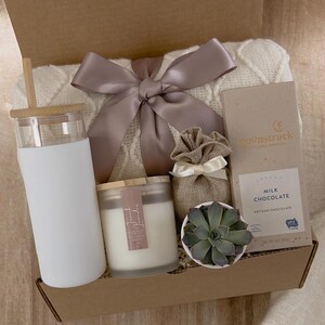 May include: A gift box filled with a white blanket, a white tumbler with a wooden lid, a white candle with a wooden lid, a small potted succulent, a burlap bag with a bow, and a bar of milk chocolate. The chocolate bar is wrapped in blue and white paper with the text "Gourmetstruck" and "Milk Chocolate" printed on it.