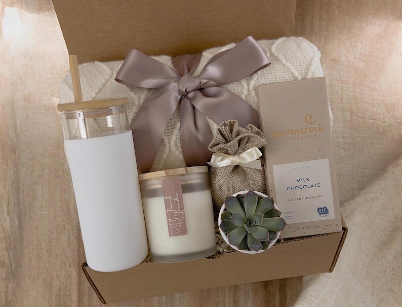 May include: A gift box filled with a white blanket, a white tumbler with a wooden lid, a white candle with a wooden lid, a small potted succulent, a burlap bag with a bow, and a bar of milk chocolate. The chocolate bar is wrapped in blue and white paper with the text "Gourmetstruck" and "Milk Chocolate" printed on it.