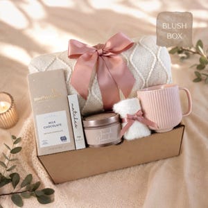 May include: A gift box containing a cream-colored knit blanket tied with a dusty pink satin ribbon, a Moonstruck milk chocolate bar, a box of matches, a French Lavender candle, a small white fuzzy item with a pink ribbon, and a ribbed pink mug.