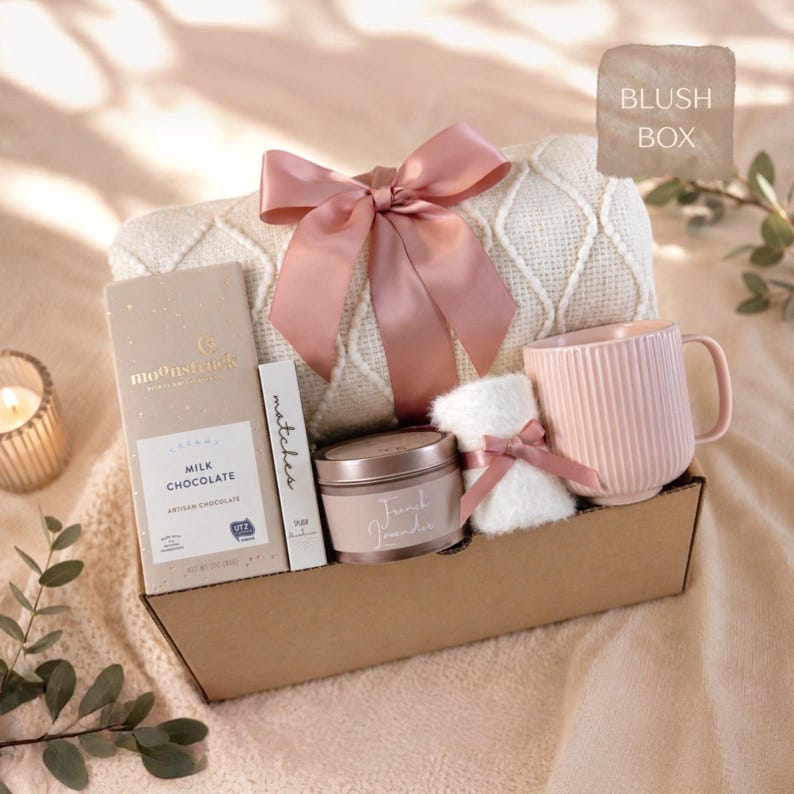 May include: A gift box containing a cream-colored knit blanket tied with a dusty pink satin ribbon, a Moonstruck milk chocolate bar, a box of matches, a French Lavender candle, a small white fuzzy item with a pink ribbon, and a ribbed pink mug.