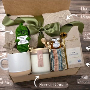 May include: A gift box filled with self-care items, including a crocheted pickle, a ceramic speckled mug, a scented candle, a blanket, gourmet chocolate, honey with a dipper, and a beverage. The box is decorated with a green ribbon and a gift tag that says "Seasonal Support Pickle".
