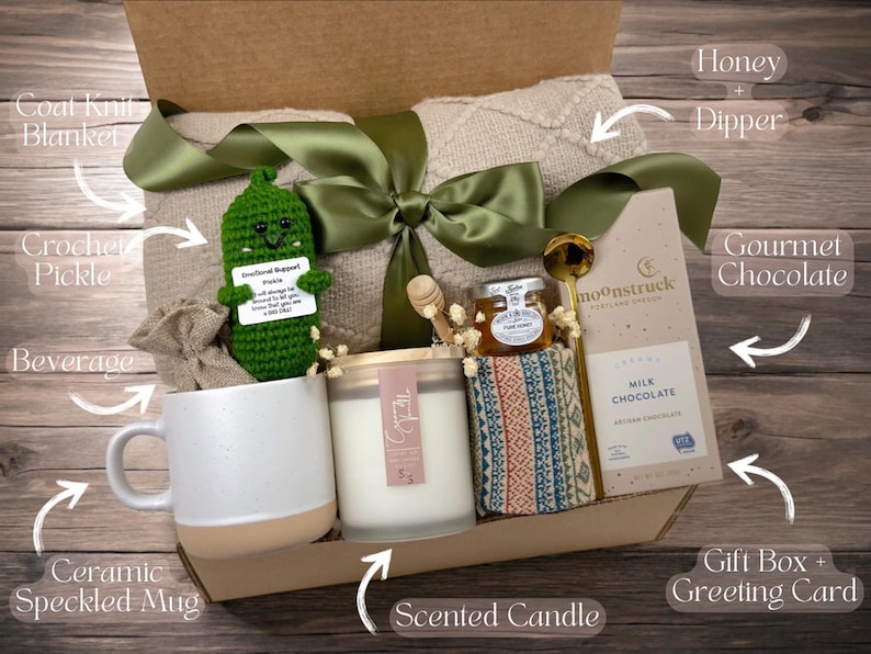 May include: A gift box filled with self-care items, including a crocheted pickle, a ceramic speckled mug, a scented candle, a blanket, gourmet chocolate, honey with a dipper, and a beverage. The box is decorated with a green ribbon and a gift tag that says "Seasonal Support Pickle".