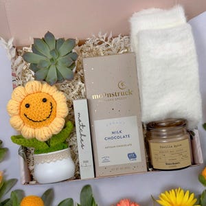 May include: A gift box filled with a variety of items, including a crocheted sunflower, a small succulent plant, a bar of milk chocolate, a candle, and a pair of fuzzy white socks. The box is lined with shredded paper and has a light pink background.