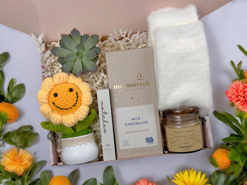 May include: A gift box filled with a variety of items, including a crocheted sunflower, a small succulent plant, a bar of milk chocolate, a candle, and a pair of fuzzy white socks. The box is lined with shredded paper and has a light pink background.