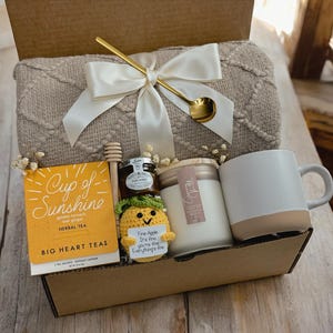 May include: A gift basket filled with a beige throw blanket, a mug, a candle, a jar of honey, a box of herbal tea, and a small crocheted pineapple with a tag that reads "Five-Apple I'm fine you're fine Everything's fine".