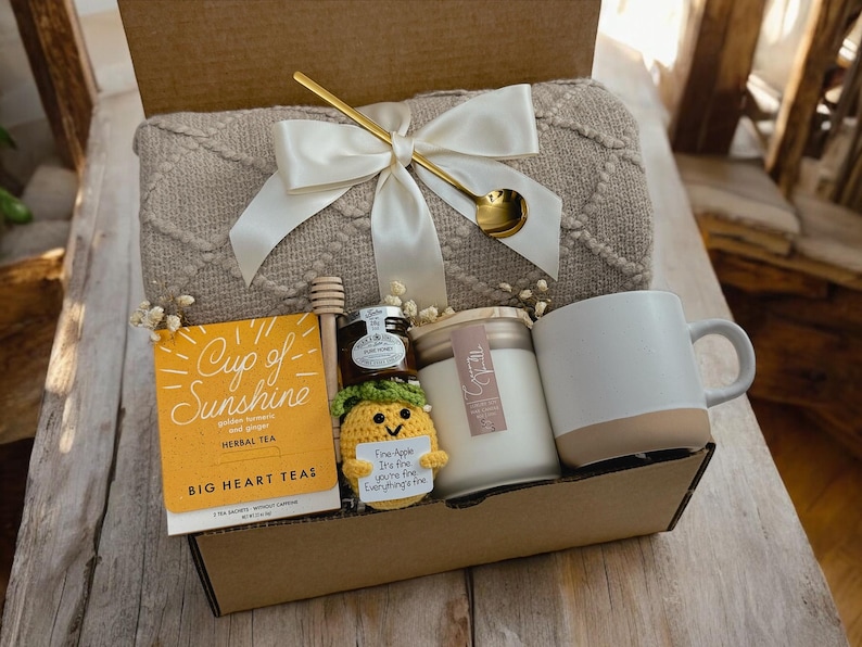 May include: A gift basket filled with a beige throw blanket, a mug, a candle, a jar of honey, a box of herbal tea, and a small crocheted pineapple with a tag that reads "Five-Apple I'm fine you're fine Everything's fine".