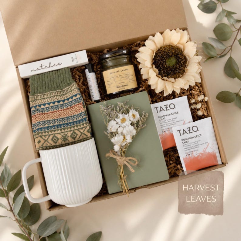 May include: A gift box containing a white ribbed mug, patterned green socks, a box of matches, a lip balm, a candle labeled Harvest Leaves, a large cream sunflower, a green notebook with dried flowers, and two Tazo Pumpkin Spice Chai tea bags.