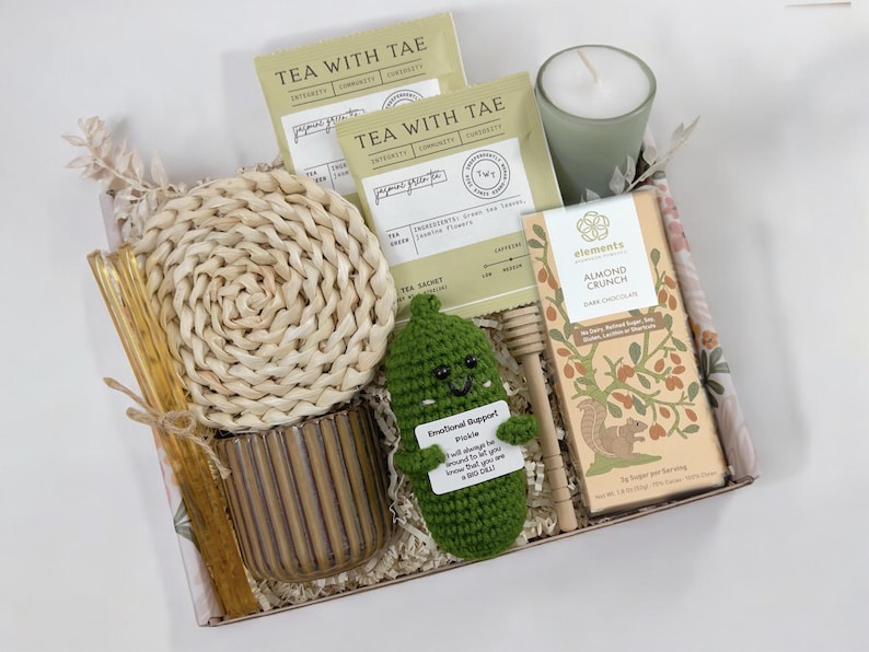 May include: A gift box filled with various items, including tea bags, a candle, chocolate, a crocheted pickle, and a wooden honey dipper. The box is lined with a floral patterned fabric. The pickle has a sign that reads "Emotional Support Pickle. I will always be there for you, no matter how bad the day is. Big Dill."