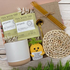 May include: A gift box filled with a white mug, a crocheted pineapple toy, two tea sachets, a gold spoon, a woven coaster, and a straw. The tea sachets are labeled "Tea with Tae" and the pineapple toy has a tag that reads "Fine-Apple It's fine you're fine Everything's fine."