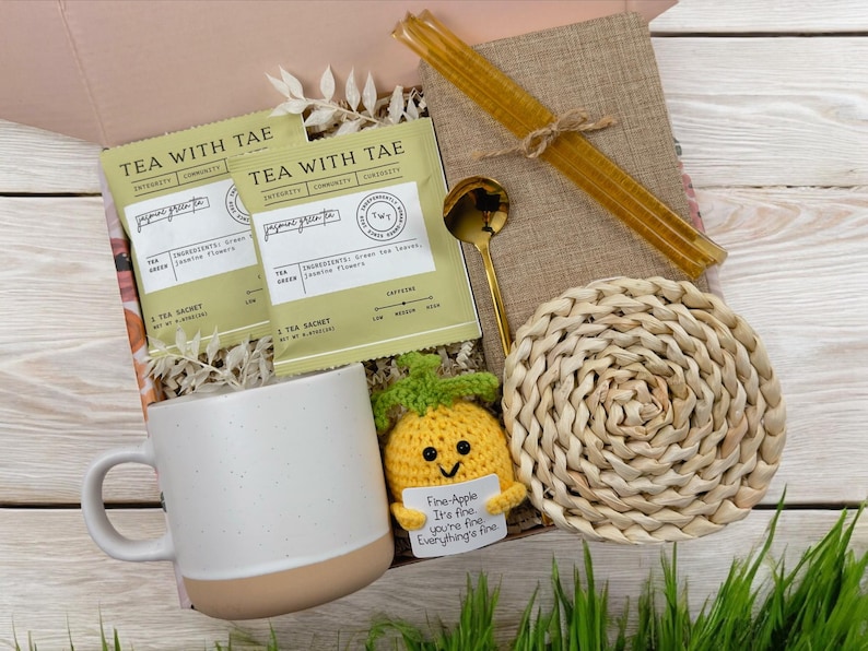 May include: A gift box filled with a white mug, a crocheted pineapple toy, two tea sachets, a gold spoon, a woven coaster, and a straw. The tea sachets are labeled "Tea with Tae" and the pineapple toy has a tag that reads "Fine-Apple It's fine you're fine Everything's fine."