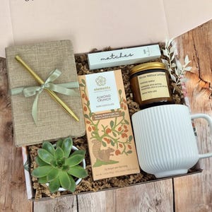 K&ouml;nnte beinhalten: Eine Geschenkbox gef&uuml;llt mit verschiedenen Artikeln, darunter ein Notizbuch, ein Stift, eine Tafel dunkle Schokolade mit Mandeln, eine Kerze, eine Tasse und eine kleine Sukkulente. Die Box ist mit braunem Papier ausgekleidet und hat einen hellrosa Deckel.