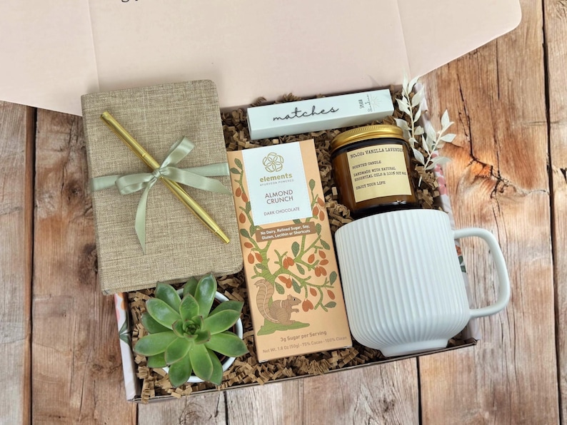K&ouml;nnte beinhalten: Eine Geschenkbox gef&uuml;llt mit verschiedenen Artikeln, darunter ein Notizbuch, ein Stift, eine Tafel dunkle Schokolade mit Mandeln, eine Kerze, eine Tasse und eine kleine Sukkulente. Die Box ist mit braunem Papier ausgekleidet und hat einen hellrosa Deckel.