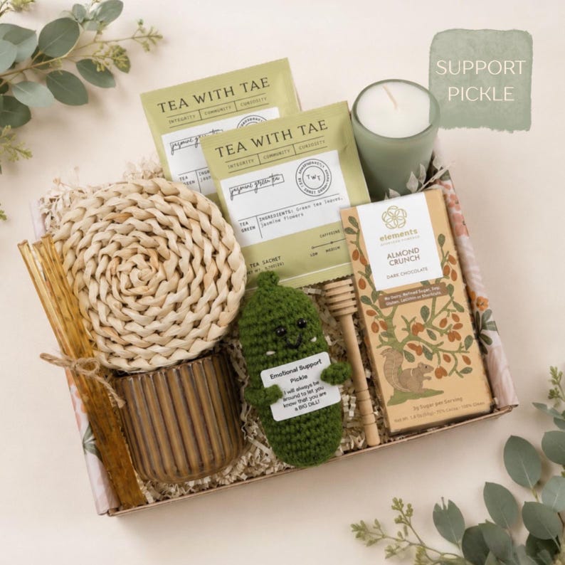K&ouml;nnte beinhalten: Eine Geschenkbox mit zwei Tea With Tae Jasmin-Gr&uuml;ntee-Beuteln, einer gr&uuml;nen Kerze im Glas, einem runden geflochtenen Untersetzer, einem braunen gerippten Keramikbecher, einem Honigl&ouml;ffel aus Holz und einer Tafel Elements Mandel-Crunch-Zartbitterschokolade mit Eichh&ouml;rnchen-Illustration. Ein kleiner gr&uuml;ner geh&auml;kelter G&uuml;rkchen mit einem Schild mit der Aufschrift Emotional Support Pickle ist enthalten.