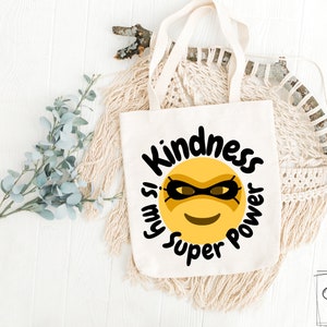 Puede incluir: Bolsa de tela blanca con una cara sonriente amarilla que lleva una máscara negra y el texto "La amabilidad es mi superpoder".