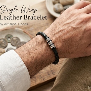 Peut inclure: Un bracelet en cuir tressé noir doté de trois perles cylindriques en métal argenté, chacune gravée d'une lettre. Le texte "Single Wrap Leather Bracelet by Artisanal Goods" est visible dans le coin supérieur gauche de l'image.