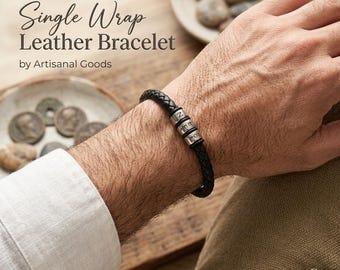 May include: A black braided leather bracelet featuring three cylindrical silver-toned metal beads, each engraved with a single letter. The text "Single Wrap Leather Bracelet by Artisanal Goods" is visible in the upper left corner of the image.