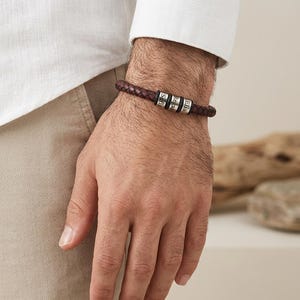 May include: A dark brown braided leather bracelet featuring three cylindrical silver-toned metal beads. Each bead is engraved with a name, specifically Sofia, Regan, and Elia, arranged in a row along the center of the braided leather band.