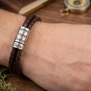 May include: A brown leather bracelet with silver name beads. The bracelet has a braided design and features the names "James", "Violet", "Evelyn", and "Chris". The bracelet is worn on a wrist.