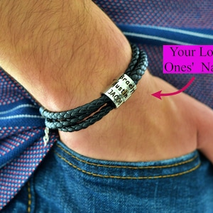 May include: Black braided leather bracelet with a silver band engraved with names. The bracelet is worn on a wrist, with the text "Your Loved Ones' Names" in a pink box in the background. The person is wearing a blue shirt and jeans.