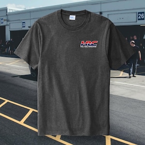 May include: Dark gray t-shirt with the HRC Honda Racing logo in red, white, and blue on the upper left chest. The shirt has a crew neck and short sleeves. The background shows a race track.