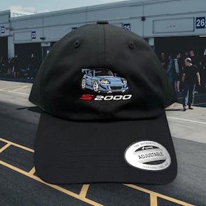 May include: Black baseball cap with an embroidered blue Honda S2000 car and the text "S2000". The cap has an adjustable strap with the text "The Authentic Yupoong Adjustable Yupoong Since 1974".