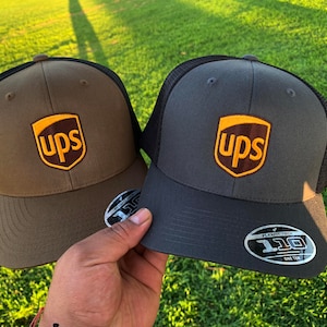 May include: Two brown and gray trucker hats with a black mesh back. Both hats have a brown UPS logo with yellow lettering on the front.