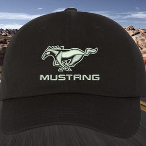 Puede incluir: Gorra de béisbol negra con el logotipo de Mustang en blanco y verde claro. El logotipo representa un caballo corriendo sobre la palabra "MUSTANG". La gorra tiene una visera curva y una corona estructurada.