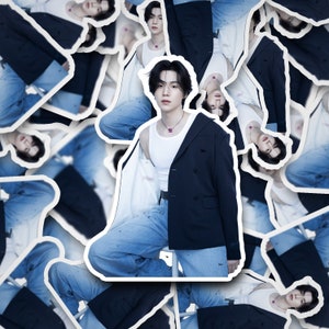 May include: A man wearing a white tank top, a dark blue blazer, and blue jeans. He is standing in front of a white background. The blazer has a distressed look and the jeans have paint splatters on them.