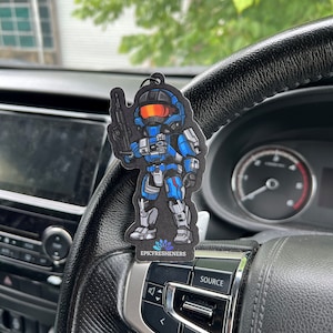Può includere: Un personaggio dei cartoni animati blu e grigio che tiene una pistola, con il testo "EPICFRESHENERS" sotto. Il personaggio è su un deodorante per auto nero.