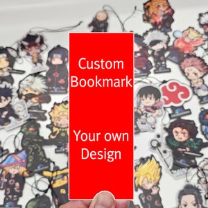 May include: A red card with the text "Custom Bookmark" and "Your own Design" on it. The card is being held by a hand in front of a white surface with many colourful cartoon character stickers.