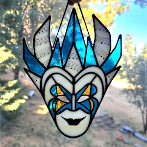 May include: A stained glass suncatcher shaped like a theatrical mask, featuring white, blue, and yellow glass panels. The upper sections incorporate translucent glass printed with black musical notation, while the lower face displays a smiling expression.