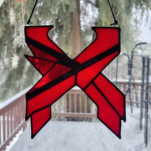 May include: A red stained glass window hanging in front of a snowy window. The window is shaped like the letter X and has black lines throughout.